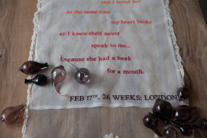 Installation art close-up of feminine lace colorfully embroidered with a poem laid on top of a rustic farm table with accents of hand blown glass filled with sand.
