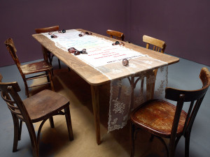 Installation art: feminine lace colorfully embroidered with a poem laid on top of a rustic farm table with accents of hand blown glass filled with sand.